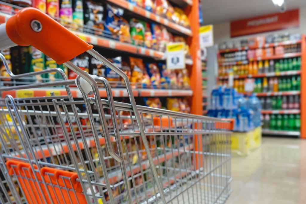 A shopping cart with an orange handle, positioned in an aisle filled with various snacks and drinks on shelves in a grocery store.